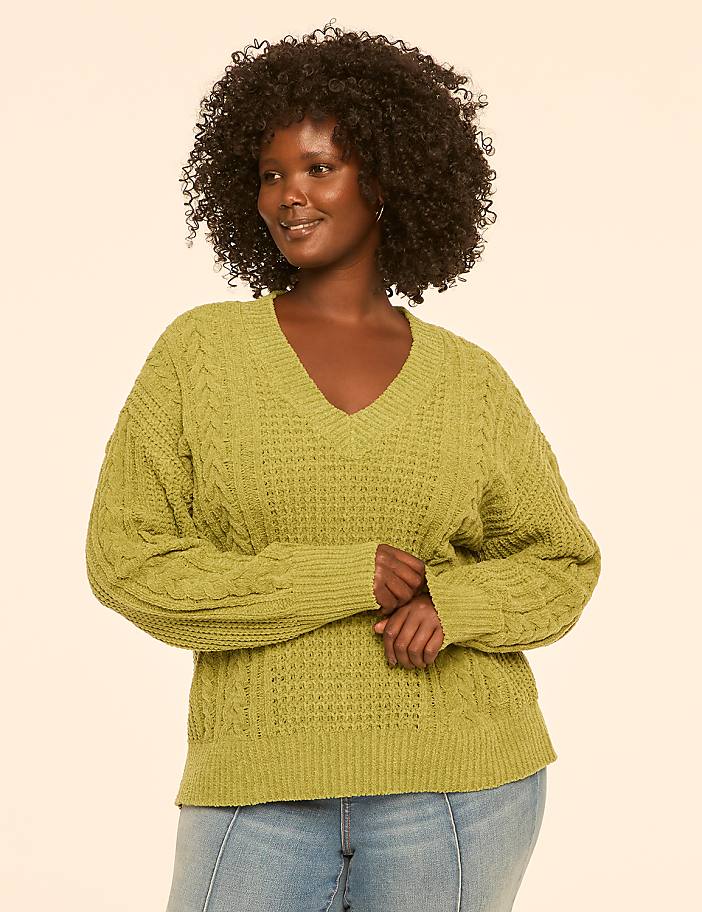 A woman with dark, curly hair and a v-neck green sweater stands against a plain, light-colored background.