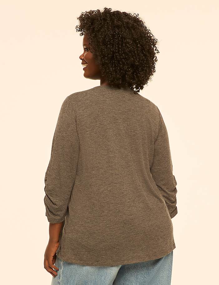 A woman with curly dark hair, wearing a grey cardigan and blue jeans, smiles over her shoulder against a plain cream background.