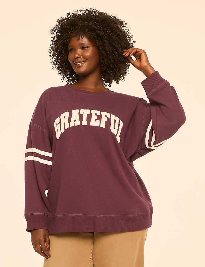 A woman with curly dark hair smiles as she poses in a maroon sweatshirt with "GRATEFUL" written across the chest, against a plain, light-colored background.