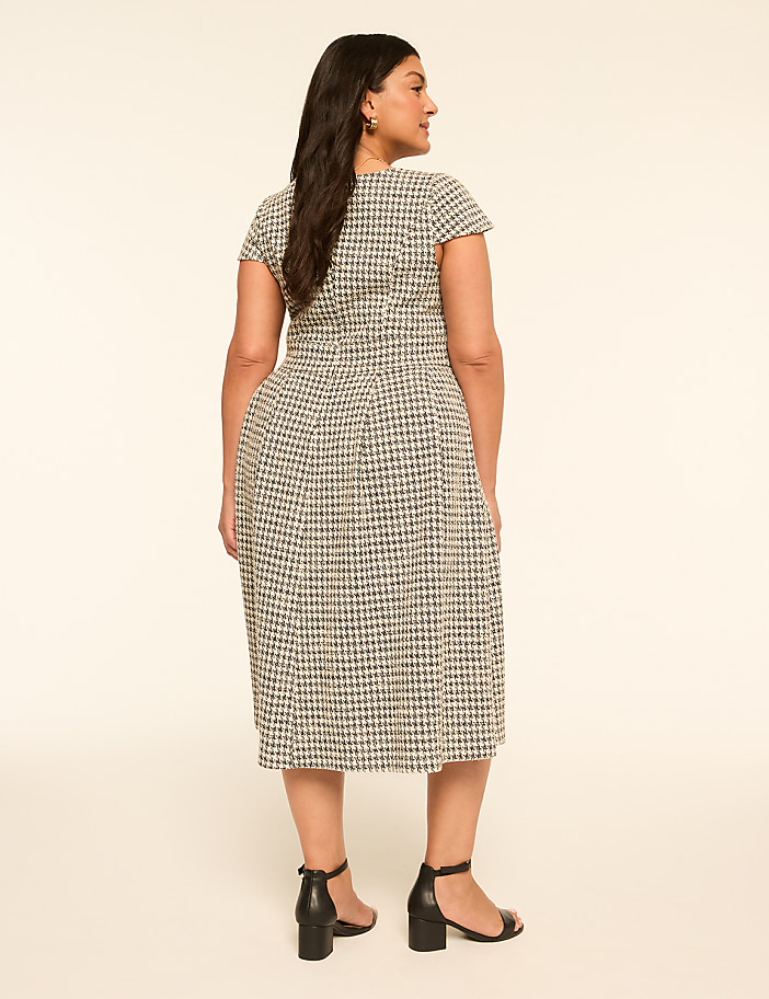 A woman in a patterned dress stands against a plain, light-colored background.