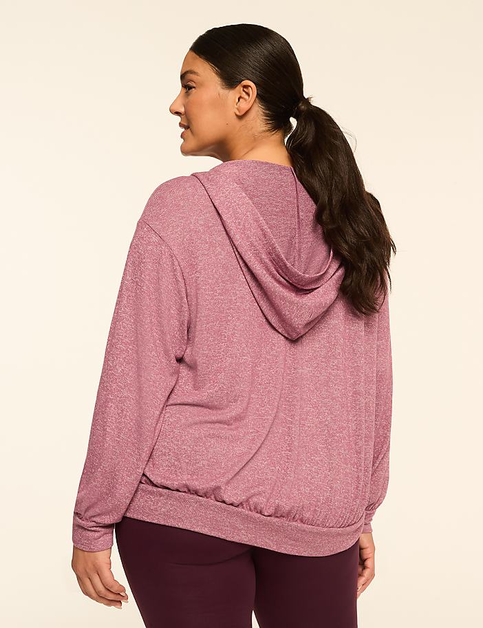 A woman in a pink hoodie and dark leggings stands against a plain, light-colored background.