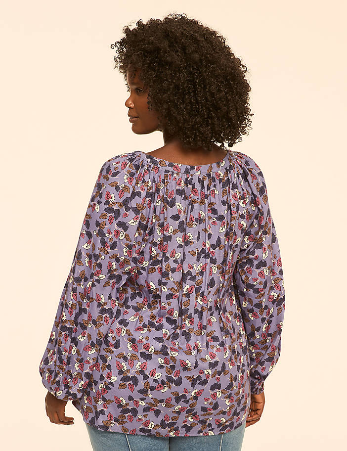 A woman with curly dark hair wears a long-sleeved, purple, floral-patterned blouse and jeans, standing against a plain, light-colored background.