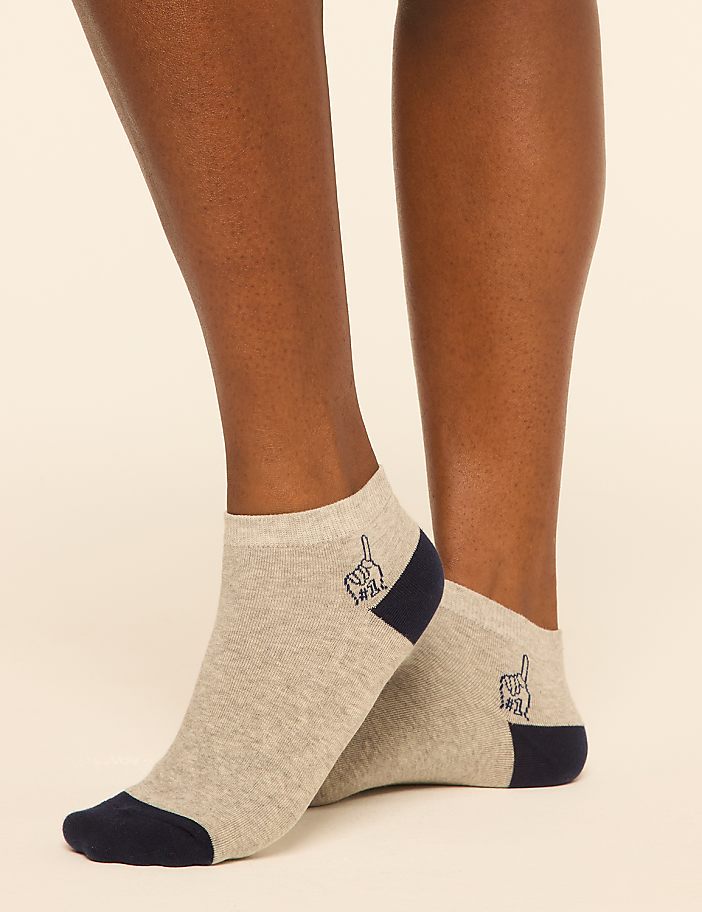 A pair of light gray ankle socks with a navy blue heel and toe, featuring a small embroidered blue cat, are worn on dark-skinned legs against a plain, light background.