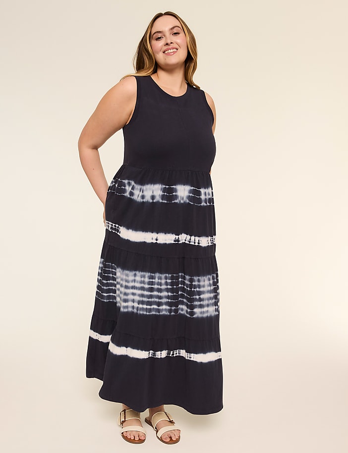 A smiling woman in a navy and white tie-dye tiered maxi dress stands against a plain, light-colored background.