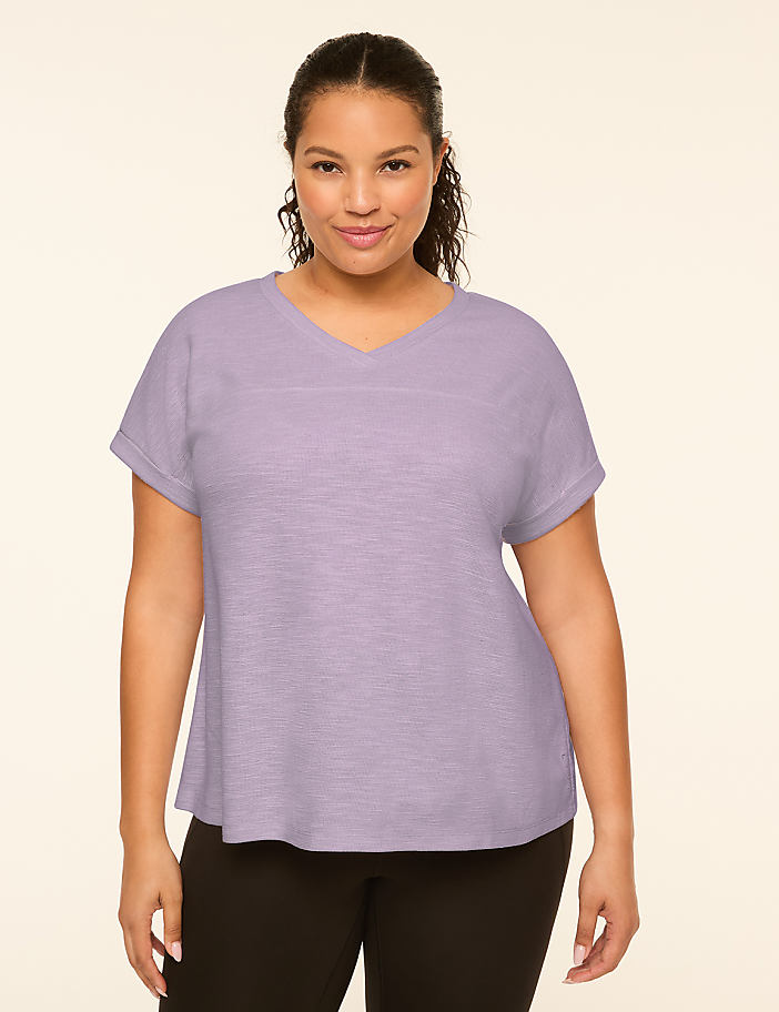A woman with dark hair and a v-neck shirt stands against a plain, light-colored background.