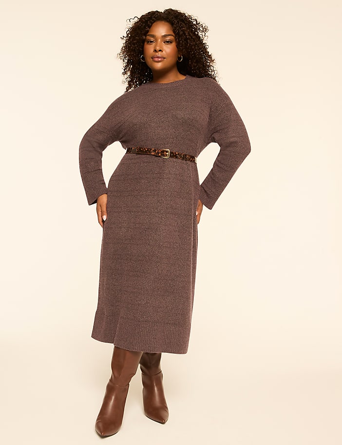 A woman in a brown knit dress and boots stands against a plain, light-colored background.