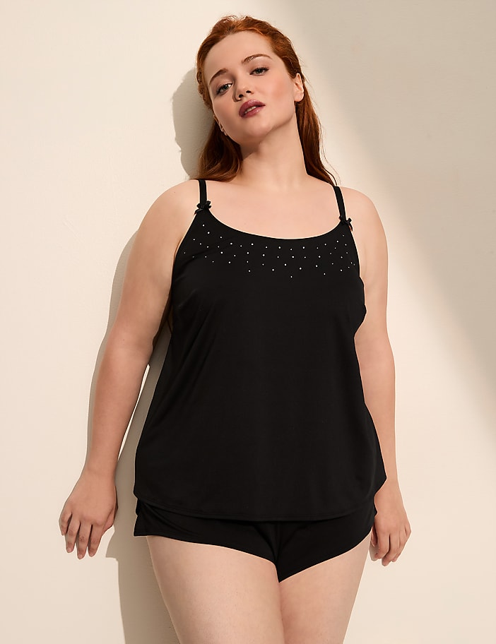 A woman in a black camisole and shorts leans against a plain, light-colored wall.