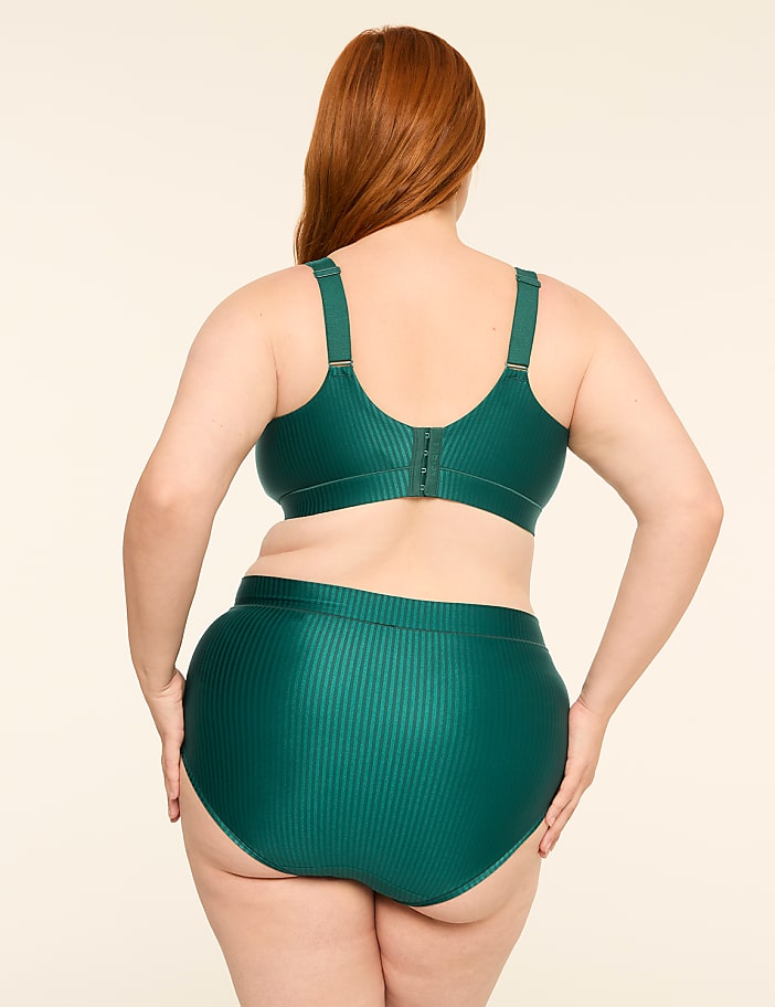 A woman with red hair stands with her back to the camera, wearing a green ribbed bra and high-waisted underwear against a plain, light-colored background.