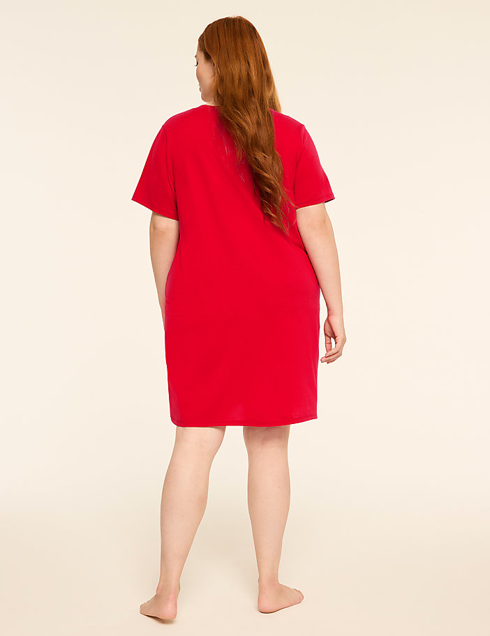 A woman with long red hair stands facing away from the camera in a red t-shirt dress against a plain, light-colored background.
