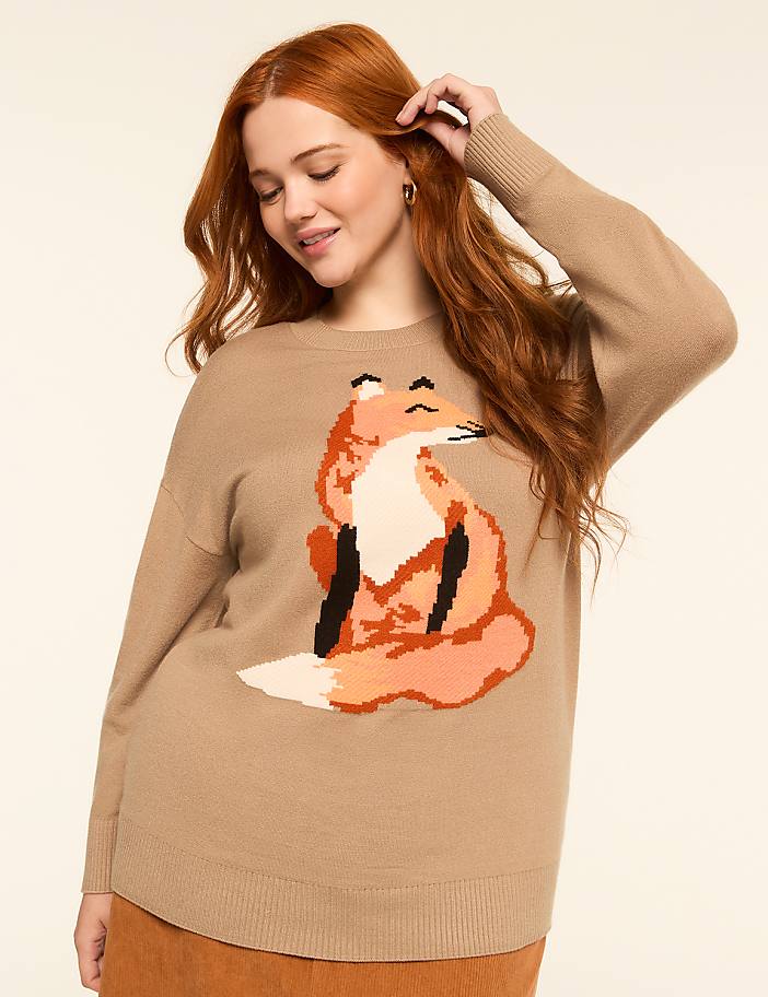 A woman with red hair smiles as she poses in a tan sweater with a pixelated fox design, against a plain cream background.