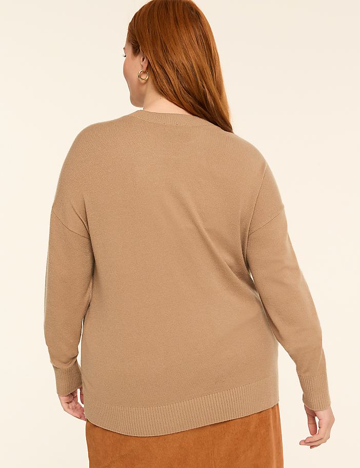 A woman with auburn hair, wearing a tan sweater and an orange skirt, stands against a plain beige background.