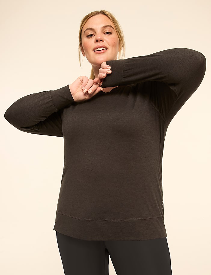 A woman in a dark long-sleeved shirt and plaid pants stretches her arms in front of a plain, light-colored background.