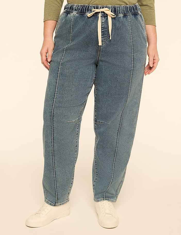 A person stands in the foreground wearing loose-fitting denim pants with a drawstring and white sneakers, against a plain, light-colored background.