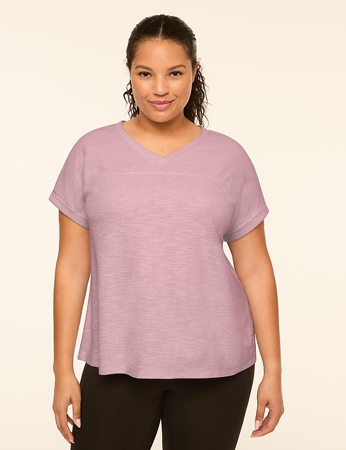 A woman with dark hair and a v-neck shirt stands against a plain, light-colored background.