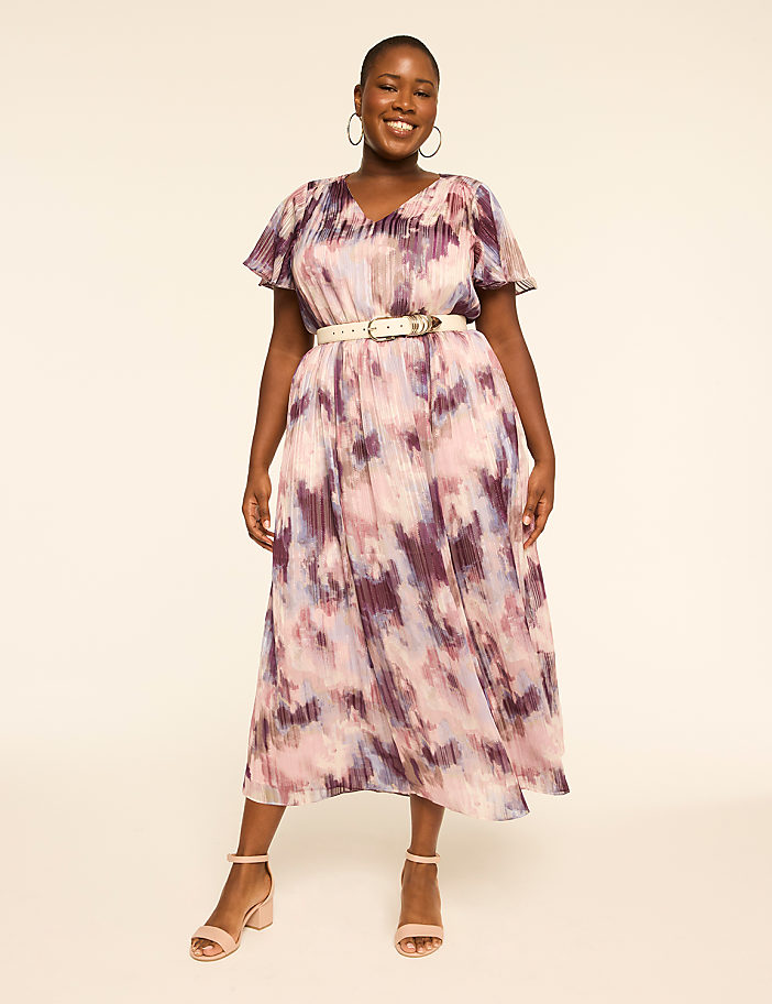 A smiling woman in a flowing, abstract-print dress and light-colored belt stands against a plain, light-colored background.