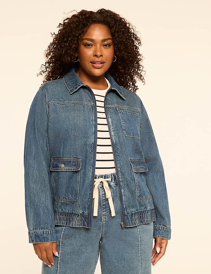 A woman in a denim jacket and striped shirt stands against a plain, light-colored background.
