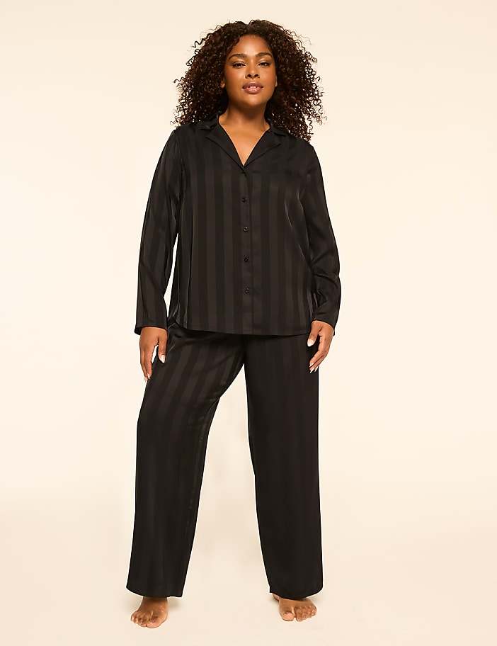 A woman with curly brown hair stands in black striped pajamas against a plain, light-colored background.