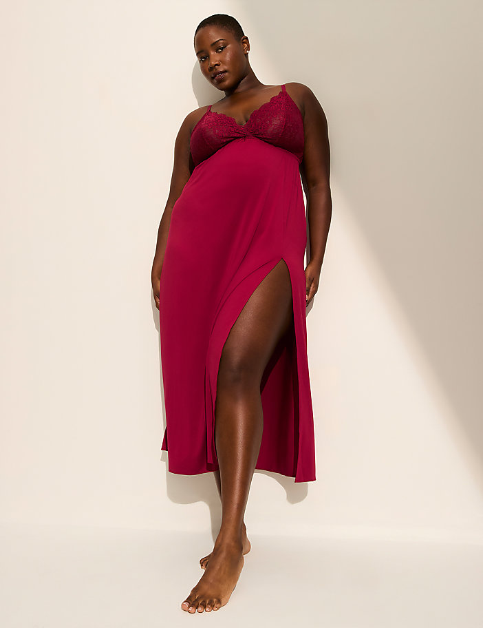 A woman in a burgundy slip dress with a thigh-high slit stands against a plain white background with soft lighting.