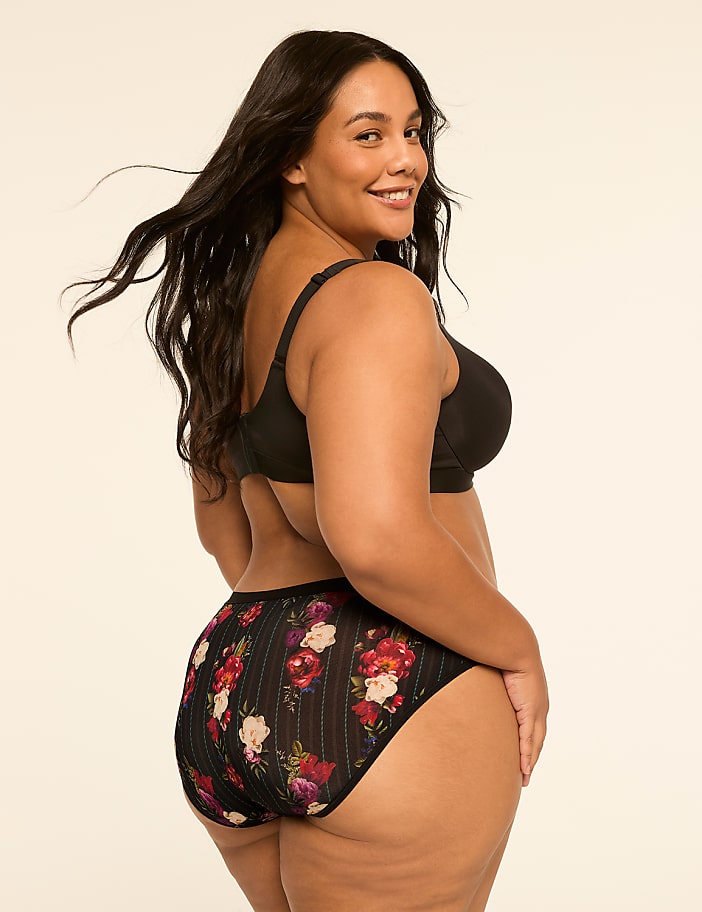 A woman with dark hair and a floral-patterned bottom is smiling over her shoulder against a plain, light-colored background.