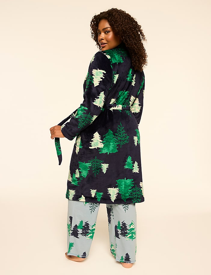A woman in a festive tree-patterned robe and pajama pants stands against a plain, light-colored background.