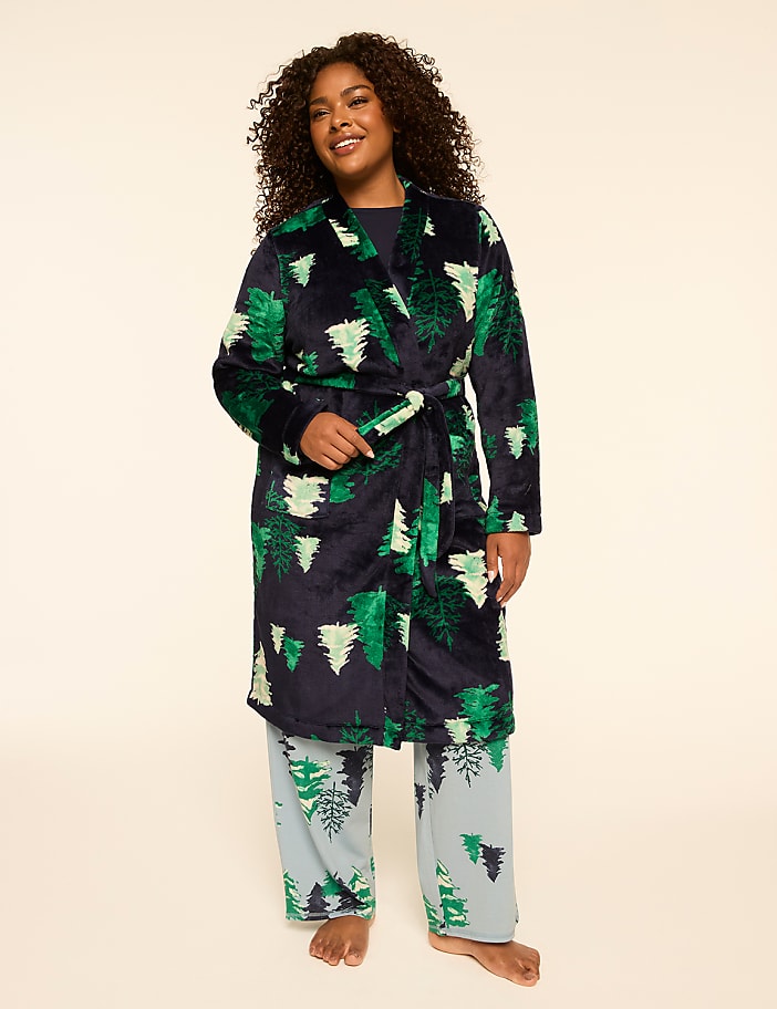 A woman in a festive green tree-patterned robe and matching pajama pants stands against a plain, light-colored background.