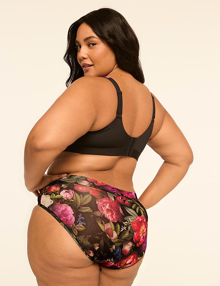 A woman in a black bra and floral-patterned underwear stands against a plain, light-colored background.