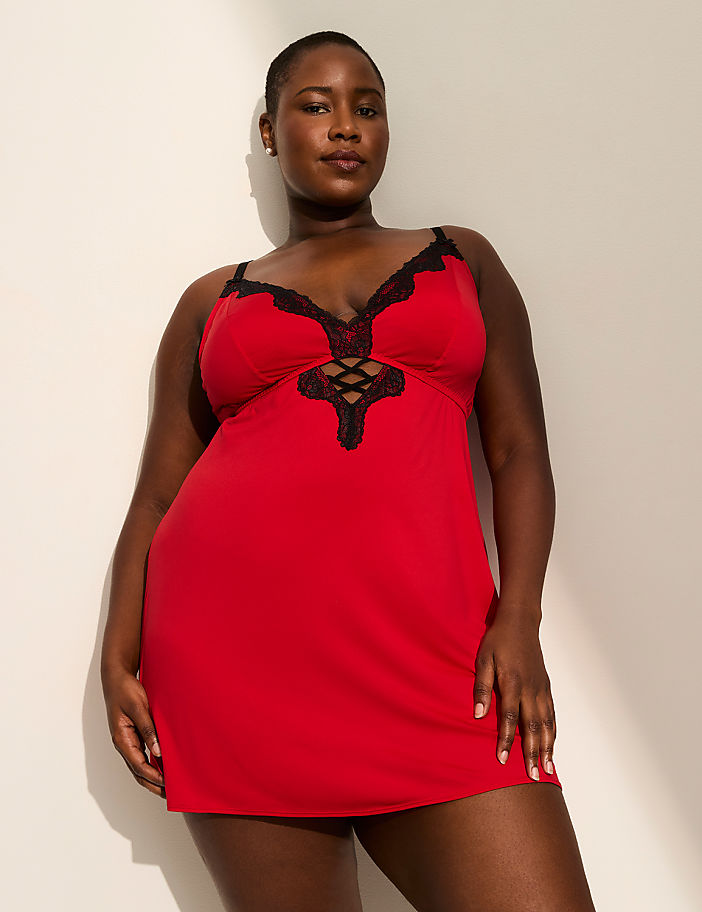 A woman in a red lace-trimmed chemise stands against a plain, light-colored wall with soft shadows.