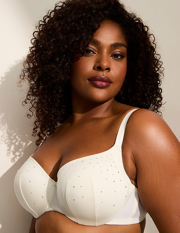 A woman with dark curly hair and makeup poses in a white bra adorned with rhinestones against a plain, light-colored background.