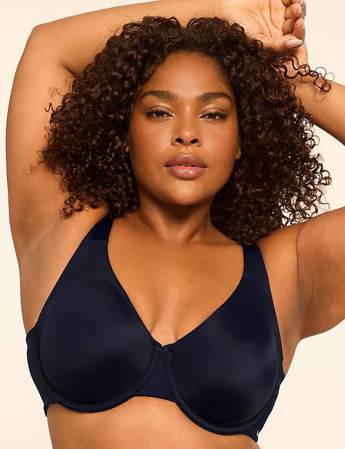 A woman with curly dark hair poses in a navy blue bra against a plain, light-colored background.