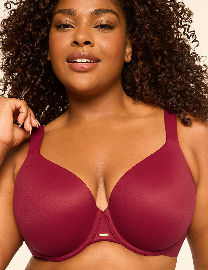A woman with curly hair smiles as she adjusts the strap of her maroon bra against a plain, light-colored background.