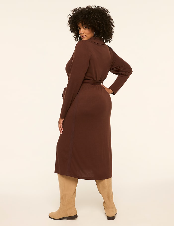 A woman in a brown dress and tan boots stands against a plain, light-colored background.
