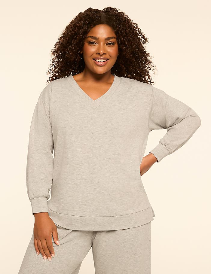 A smiling woman with curly dark hair poses in a light gray v-neck sweatshirt and matching pants against a plain, light-colored background.