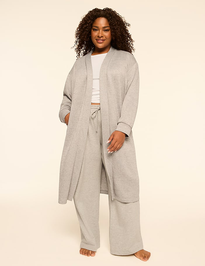 A woman in a grey robe and pants stands against a plain, light-colored background.