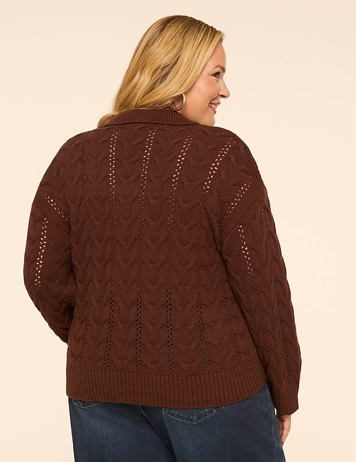 A woman in a brown cable-knit sweater and blue jeans stands against a plain, light-colored background.
