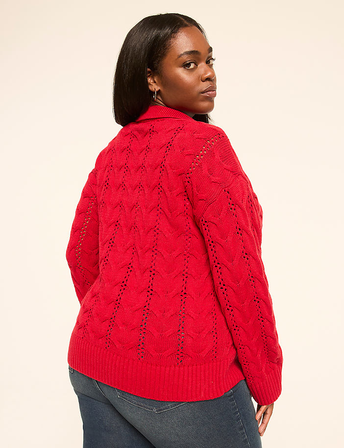 A woman in a red cable-knit sweater and blue jeans stands against a plain, light-colored background.