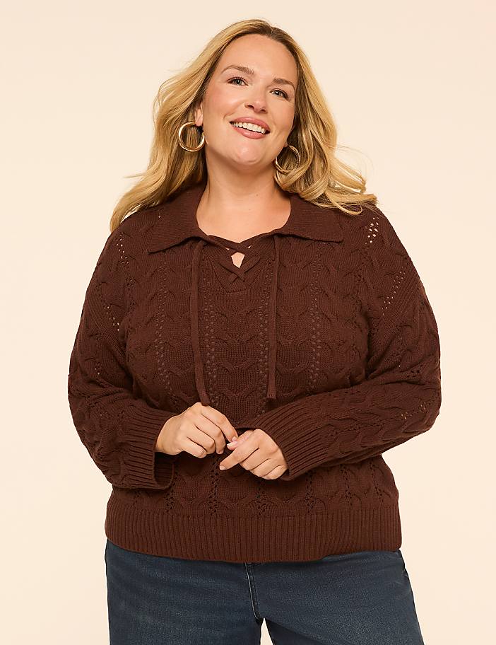 A smiling woman in a brown cable-knit sweater with a lace-up collar stands against a plain, light-colored background.