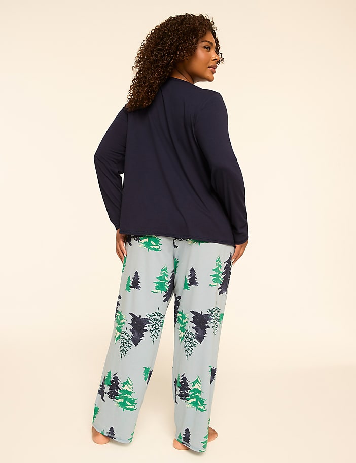 A woman in a navy blue long-sleeved top and light blue pajama pants with a green and navy tree print stands against a plain, light-colored background.