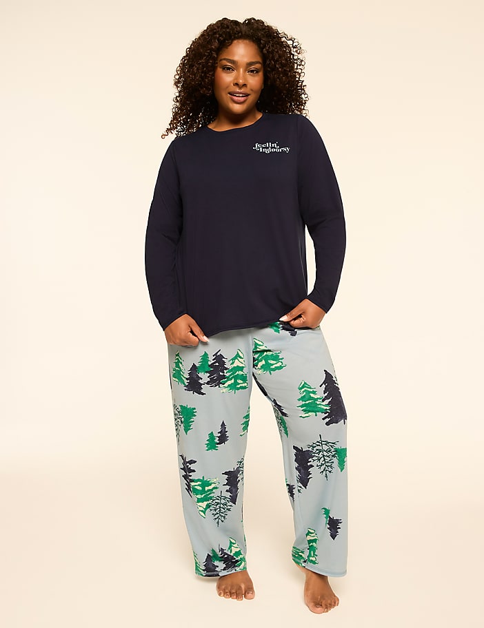 A woman stands in the foreground wearing a navy blue long-sleeved shirt and light blue pajama pants with a forest print, against a plain, light-colored background.
