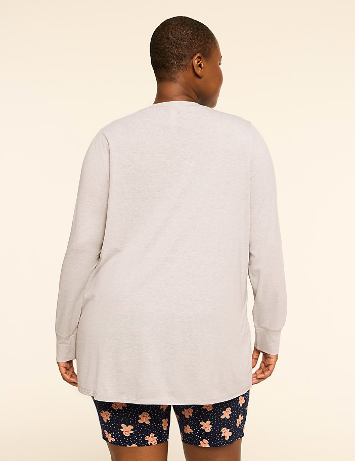 A person wearing a light gray long-sleeved shirt and patterned shorts stands against a plain, light-colored background.