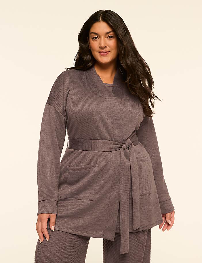 A woman in a grey waffle-knit robe with a tie belt stands against a plain, light-colored background.