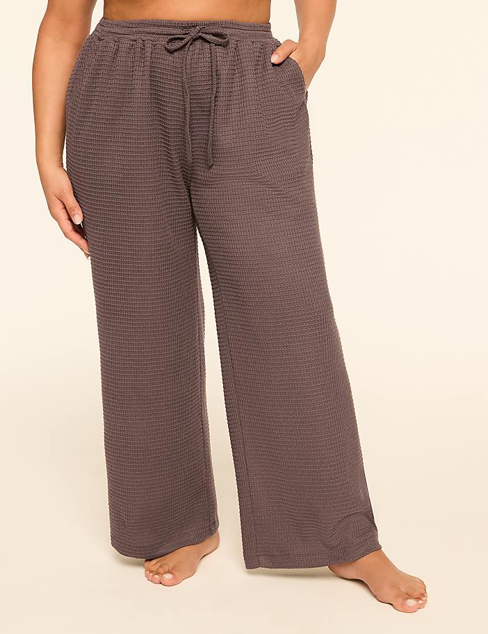 A person stands in the foreground wearing loose-fitting, waffle-knit, charcoal-colored pajama pants with a drawstring, against a plain, light beige background.