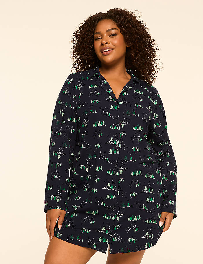 A woman with curly brown hair smiles at the camera, wearing a dark blue pajama set with a forest print, against a plain beige background.