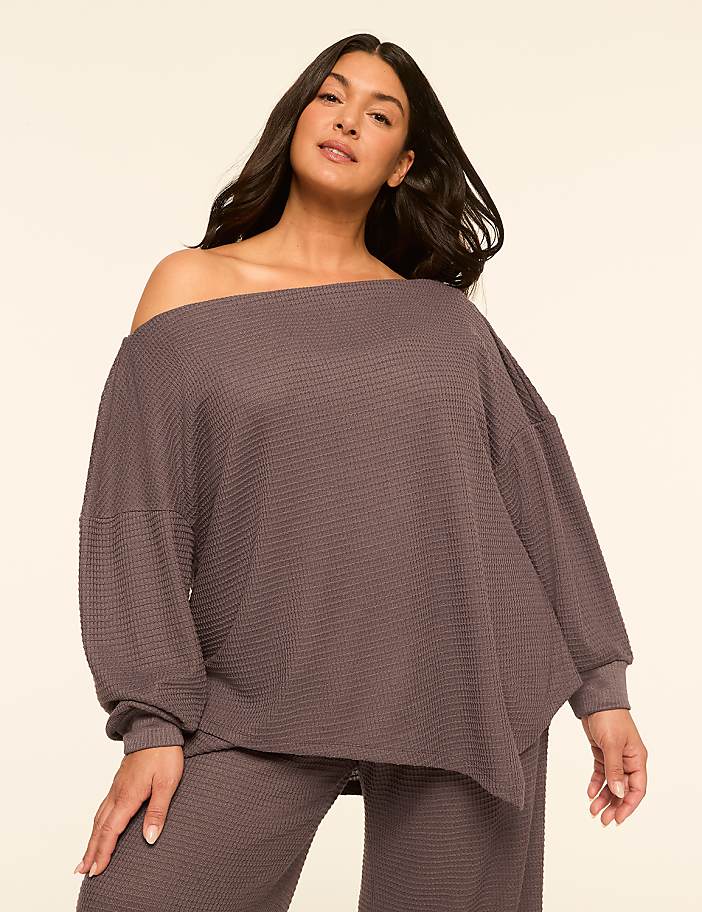 A woman in a waffle-knit off-the-shoulder top and matching pants poses against a plain, light-colored background.