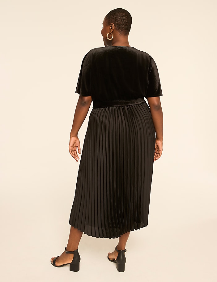A woman in a black velvet top and pleated skirt stands against a plain, light-colored background.