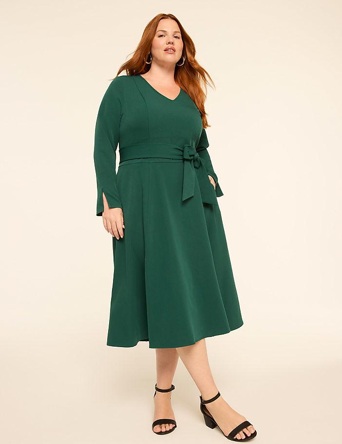 A woman in a green dress stands against a plain, light-colored background.