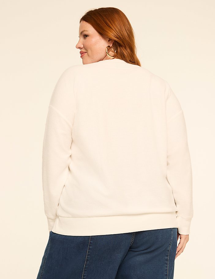 A woman with red hair wearing a cream-colored sweatshirt and blue jeans stands against a plain, light-colored background.
