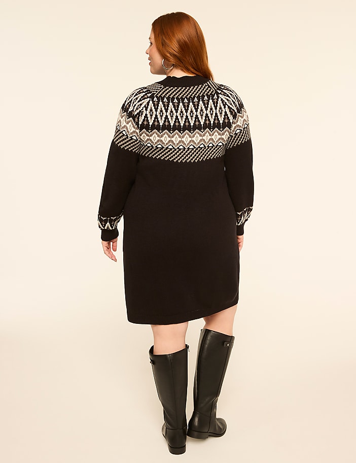 A woman in a black dress with a fair isle pattern on the yoke and black boots stands against a plain, light-colored background.
