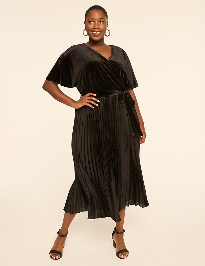 A smiling woman in a black velvet wrap dress with a pleated skirt stands against a plain, light-colored background.