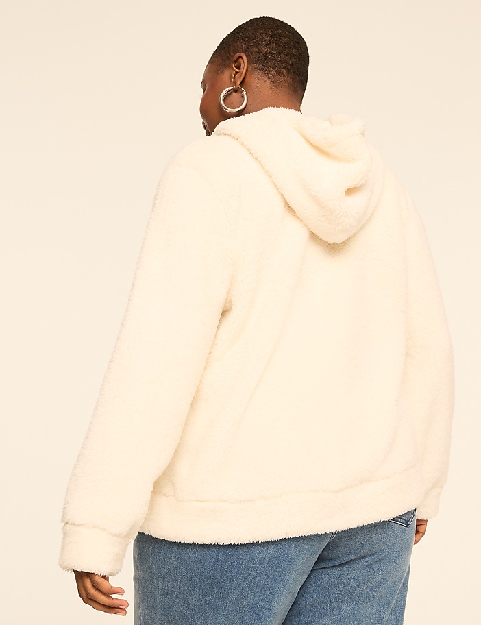 A person in a cream-colored fuzzy hoodie and blue jeans stands against a plain, light-colored background.