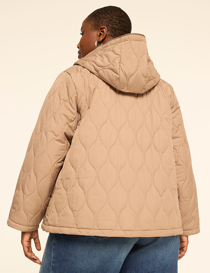 A woman in a tan quilted jacket and blue jeans stands against a plain, light-colored background.